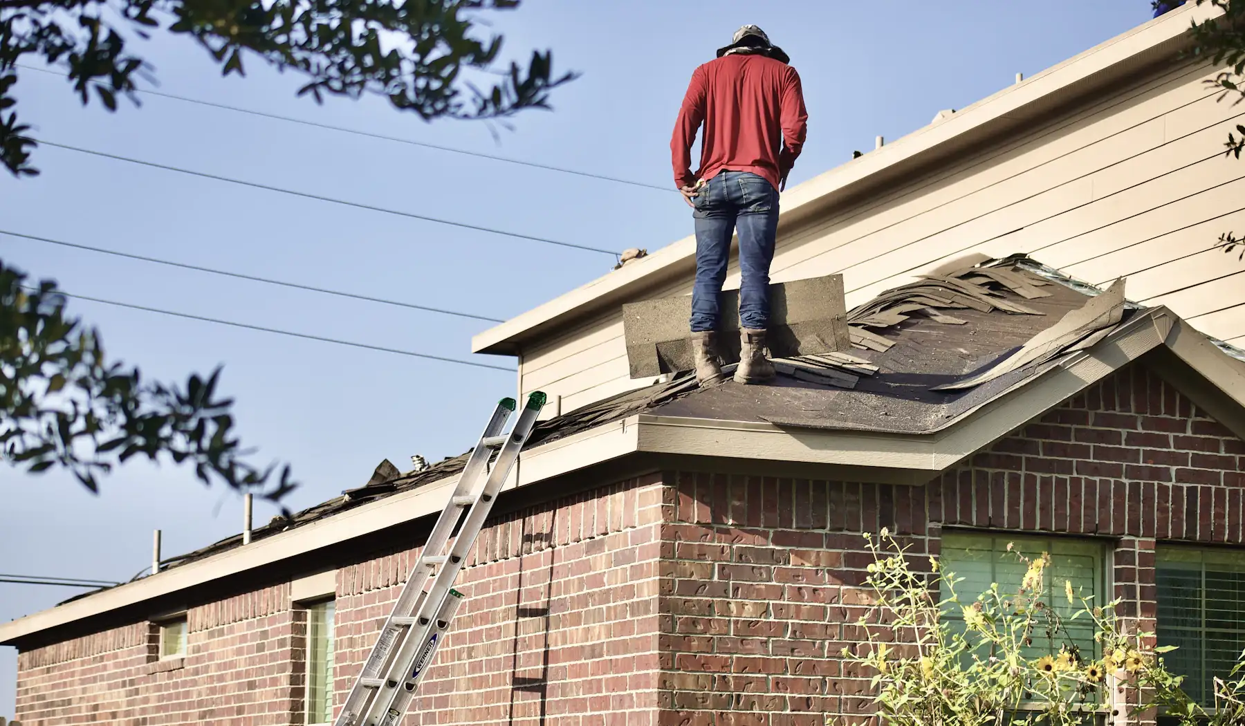 A person standing on a house roof with worn shingles, a ladder against the brick wall below